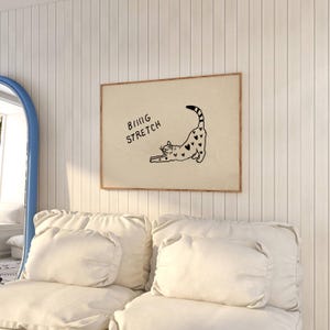 Puede incluir: Un estampado enmarcado con un gato negro estir&aacute;ndose con marcas en forma de coraz&oacute;n. Las palabras "BIG STRETCH" est&aacute;n encima del gato. El estampado tiene un fondo beige y un marco marr&oacute;n, colgado en una pared blanca.