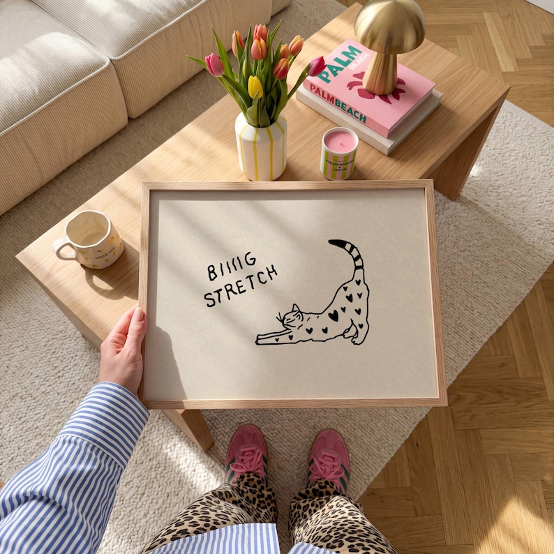 Puede incluir: Un cuadro enmarcado con un gato negro estir&aacute;ndose, adornado con patrones en forma de coraz&oacute;n. El texto "BIG STRETCH" est&aacute; encima del gato. La persona lleva una camisa a rayas azules y blancas y pantalones con estampado de leopardo.