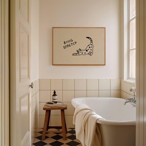 Puede incluir: En un cuarto de ba&ntilde;o, una impresi&oacute;n enmarcada muestra un gato negro estir&aacute;ndose con marcas en forma de coraz&oacute;n y las palabras "BIG STRETCH". El ba&ntilde;o tiene una ba&ntilde;era blanca, un taburete de madera y un suelo a cuadros blancos y negros.