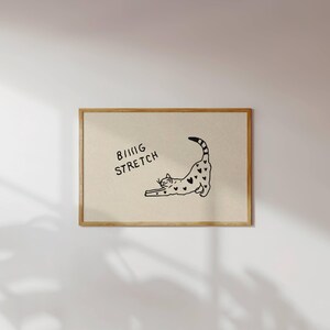 Puede incluir: Una impresi&oacute;n enmarcada con una ilustraci&oacute;n en blanco y negro de un gato estir&aacute;ndose, adornada con patrones en forma de coraz&oacute;n. Las palabras "BIG STRETCH" est&aacute;n escritas encima del gato. La obra de arte se presenta sobre un fondo neutro.