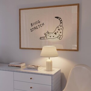 Puede incluir: Una obra de arte enmarcada con una ilustraci&oacute;n de un gato estir&aacute;ndose, con el texto "BIG STRETCH". Una l&aacute;mpara blanca con pantalla est&aacute; sobre un mueble blanco. Hay libros apilados en el mueble.