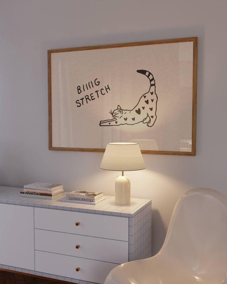 Puede incluir: Una obra de arte enmarcada con una ilustraci&oacute;n de un gato estir&aacute;ndose, con el texto "BIG STRETCH". Una l&aacute;mpara blanca con pantalla est&aacute; sobre un mueble blanco. Hay libros apilados en el mueble.