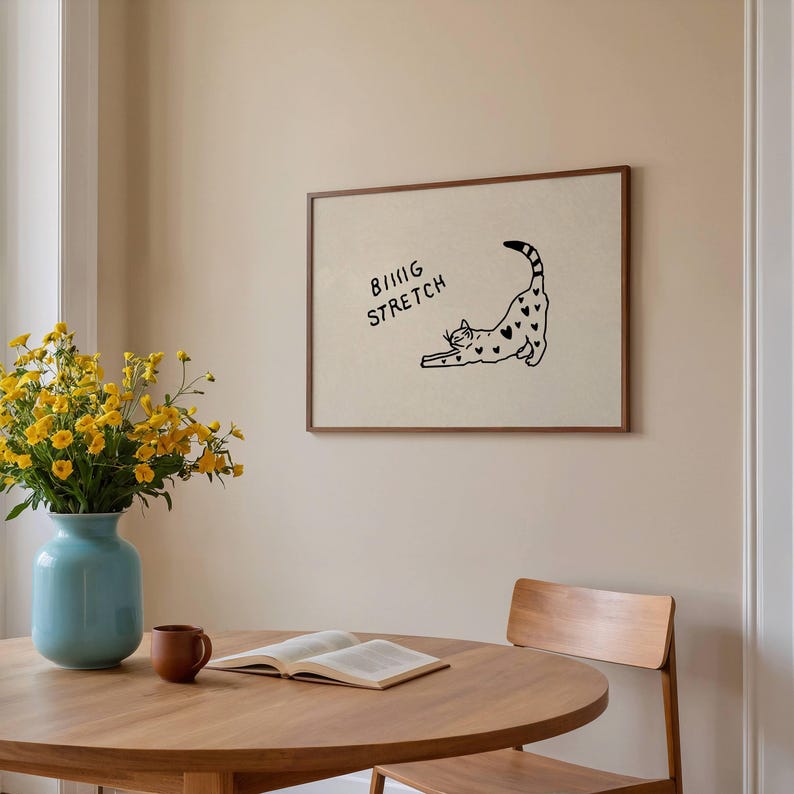 Puede incluir: Un cuadro enmarcado con un gato negro estir&aacute;ndose, con marcas en forma de coraz&oacute;n. El texto "BIG STRETCH" est&aacute; encima del gato. El cuadro est&aacute; en un marco marr&oacute;n, colgado en una pared beige. Una mesa redonda de madera, un jarr&oacute;n con flores amarillas y un libro est&aacute;n en primer plano.