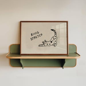 Puede incluir: Un cuadro enmarcado con una ilustraci&oacute;n de un gato negro estir&aacute;ndose, adornado con corazones. El texto "BIG STRETCH" est&aacute; encima del gato. El cuadro se muestra en una estanter&iacute;a verde y de madera.