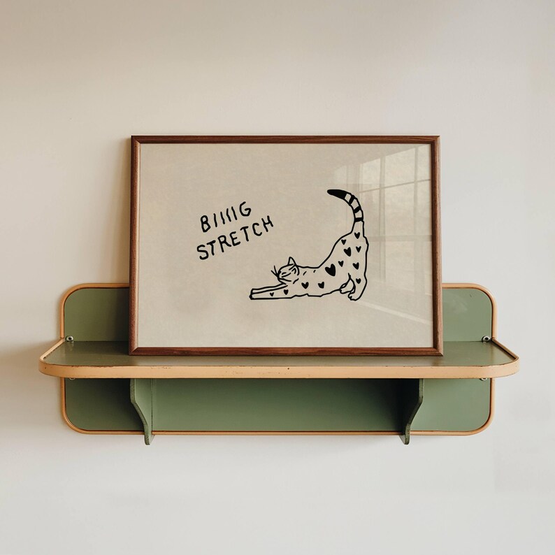 Puede incluir: Un cuadro enmarcado con una ilustraci&oacute;n de un gato negro estir&aacute;ndose, adornado con corazones. El texto "BIG STRETCH" est&aacute; encima del gato. El cuadro se muestra en una estanter&iacute;a verde y de madera.