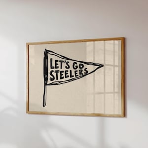 Peut inclure: Une impression d'art murale encadrée présentant une illustration de fanion dessinée à la main en noir sur un fond beige clair. Le fanion contient le texte noir en gras "LET'S GO STEELERS" écrit dans un style de police manuscrit décontracté.
