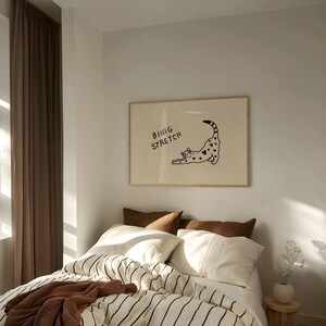 Puede incluir: Un cuadro enmarcado con una ilustraci&oacute;n de un gato negro estir&aacute;ndose, con corazones y el texto "BIG STRETCH". Sobre una cama con ropa de cama a rayas y cojines marrones.