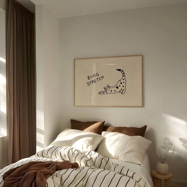 Puede incluir: Un cuadro enmarcado con una ilustraci&oacute;n de un gato negro estir&aacute;ndose, con corazones y el texto "BIG STRETCH". Sobre una cama con ropa de cama a rayas y cojines marrones.