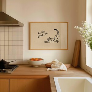 Puede incluir: Un cuadro con una ilustraci&oacute;n de un gato negro estir&aacute;ndose, adornado con corazones. El texto "BIG STRETCH" est&aacute; encima del gato. La imagen est&aacute; en una cocina con una encimera de madera, un cuenco de naranjas y un fregadero blanco.