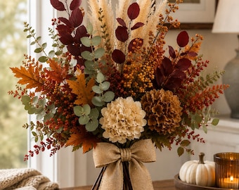 May include: A decorative floral arrangement featuring cream and brown hydrangea blossoms, tall beige pampas grass, deep red eucalyptus leaves, orange berries, and autumn-colored maple leaves, all tied together with a wide, natural burlap bow.