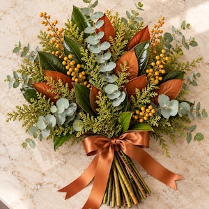 May include: A floral bouquet featuring dark green magnolia leaves, copper-toned magnolia leaves, sprigs of green cedar, silver-dollar eucalyptus stems, and clusters of small yellow berries, all tied together with a wide, smooth, copper-colored satin ribbon.