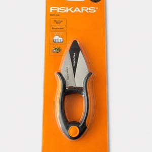 May include: Fiskars Herb Snip in orange and black packaging. The tool features stainless steel blades and a black handle with an orange circular button. The packaging includes the text Fiskars, Herb Snip, Stainless steel, Easy Action, and Full Lifetime Warranty.