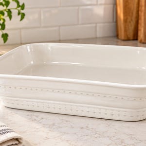 May include: A rectangular white ceramic baking dish with rounded corners and a smooth, glossy finish. The exterior walls feature two horizontal bands of embossed dot detailing that wrap around the entire perimeter of the vessel.