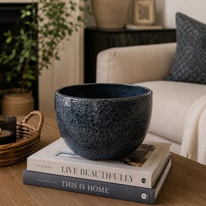 May include: A deep blue, speckled ceramic bowl with a rounded shape sits atop two stacked hardcover books. The top book is titled Live Beautifully by Calderone, and the bottom book is titled This is Home by Natalie Walton, both with white text.