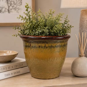 May include: A round ceramic planter with a glossy glaze that transitions from a dark brown rim to a vibrant mustard yellow and olive green base. The planter is filled with a lush, bushy artificial green plant with small, rounded leaves.