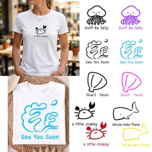 Op de afbeelding: Witte T-shirts met maritieme ontwerpen. Eén shirt heeft een krabbenafbeelding en de tekst "a little crabby". Andere ontwerpen tonen kwallen, schelpen en walvissen met zinnen als "Don't Be Jelly" en "Sea You Soon."