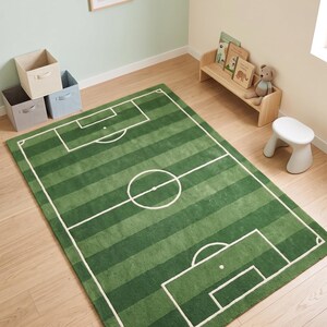 May include: A rectangular area rug featuring a green football pitch design with alternating light and dark green stripes. The rug includes white lines depicting the penalty areas, the centre circle, and the boundary lines of a football pitch.