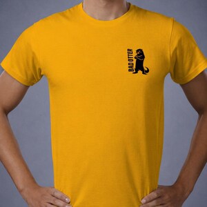May include: A mustard yellow short-sleeved t-shirt featuring a small black graphic on the left chest. The graphic depicts a silhouette of an otter standing with its arms crossed, accompanied by the vertical black text that reads BAD OTTER.