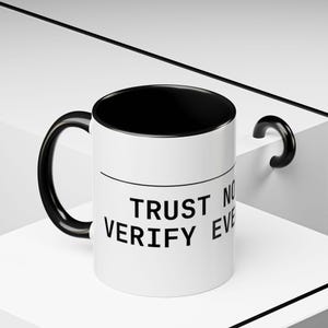May include: White ceramic mug with a black interior and handle. The mug features the text "TRUST NO ONE VERIFY EVERYTHING" in black, centered on the front. The mug is set against a white and black geometric background.
