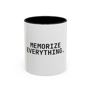 Könnte beinhalten: Weißer Keramikbecher mit schwarzem Innenraum. Der Becher trägt den Text "MEMORIZE EVERYTHING" in fetten, schwarzen Großbuchstaben. Der Becher ist zylindrisch und steht auf einer weißen Oberfläche.