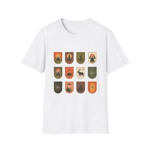 Puede incluir: Camiseta blanca con una serie de diseños gráficos inspirados en la naturaleza. Cada diseño tiene forma de escudo y presenta ilustraciones de montañas, árboles, un oso, un ciervo y una brújula. Los diseños utilizan colores naranja, marrón y verde.