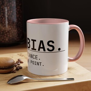 May include: White ceramic mug with a pink interior and handle. The mug features the word "BIAS" in black, along with the phrases "IMPORTANCE" and "THE POINT." A spoon and a macaron are on a wooden surface.