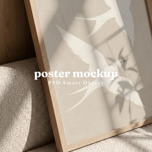 Peut inclure: Une affiche dans un cadre en bois clair représentant deux oiseaux blancs en vol sur un fond beige. Le texte By Barakaya, poster mockup et PSD Smart Object est imprimé sur la surface. L'image présente des ombres de feuilles sur le cadre.