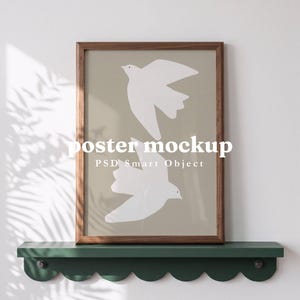 May include: A rectangular wooden frame containing a beige poster with two white bird silhouettes. The frame rests on a dark green scalloped shelf. The text "poster mockup" and "PSD Smart Object" are centered on the image, with "By Barakaya" above.