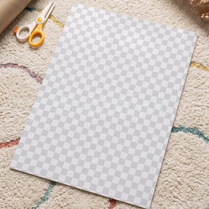 May include: A rectangular sheet of paper featuring a repeating light grey and white checkerboard pattern. The paper is positioned flat, accompanied by a pair of white and yellow scissors and a brown cardboard tube nearby.