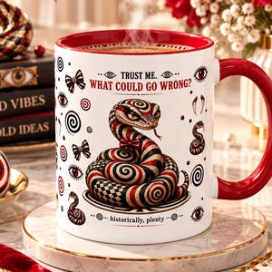 Op de afbeelding: Een witte keramische mok met een rode binnenkant en handvat, voorzien van een afbeelding van een opgerolde slang in rood, zwart en goud. De tekst luidt "TRUST ME. WHAT COULD GO WRONG?" en "historically, plenty" met kleine oog- en spiraalpictogrammen.