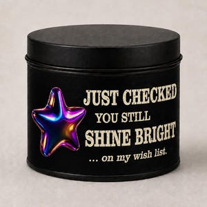 Puede incluir: Una lata de vela redonda negra con tapa. La lata presenta una estrella hologr&aacute;fica y el texto "JUST CHECKED YOU STILL SHINE BRIGHT ... on my wish list."
