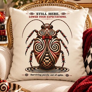 May include: A square throw pillow with a cream-colored cover featuring a central graphic of a beetle with a red and black spiral pattern and a small red bow tie. The text reads STILL HERE. LOWER YOUR EXPECTATIONS. and Surviving purely out of spite.