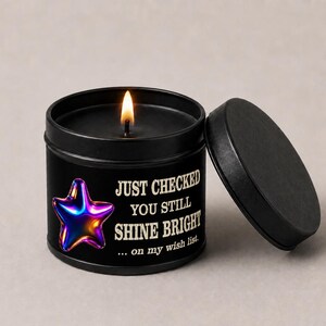 Puede incluir: Una vela negra en una lata redonda con tapa. La vela est&aacute; encendida, y la lata tiene el texto "JUST CHECKED YOU STILL SHINE BRIGHT" con un gr&aacute;fico de estrella colorida. La tapa est&aacute; a un lado.