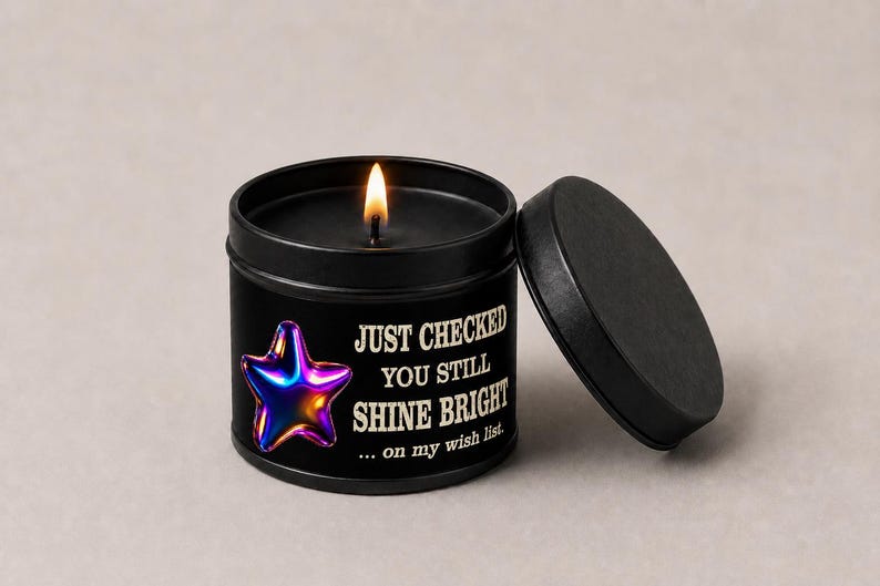 Puede incluir: Una vela negra en una lata redonda con tapa. La vela est&aacute; encendida, y la lata tiene el texto "JUST CHECKED YOU STILL SHINE BRIGHT" con un gr&aacute;fico de estrella colorida. La tapa est&aacute; a un lado.