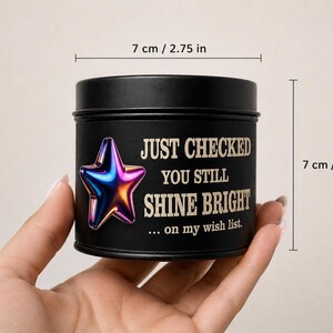 Puede incluir: Una lata de vela negra, de 7 cm de di&aacute;metro y altura, con el texto "JUST CHECKED YOU STILL SHINE BRIGHT" y un gr&aacute;fico de estrella colorida. La lata se sostiene en una mano.