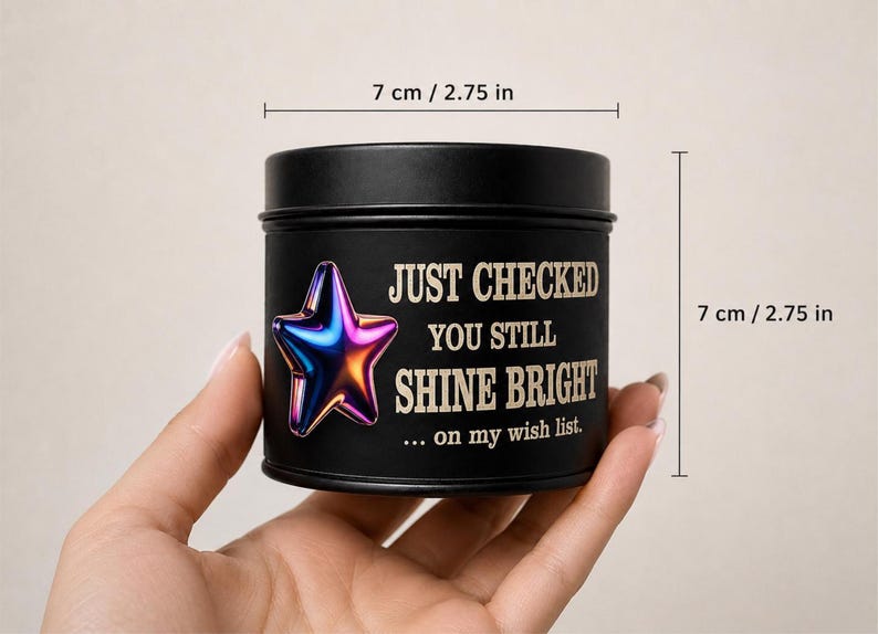 Puede incluir: Una lata de vela negra, de 7 cm de di&aacute;metro y altura, con el texto "JUST CHECKED YOU STILL SHINE BRIGHT" y un gr&aacute;fico de estrella colorida. La lata se sostiene en una mano.