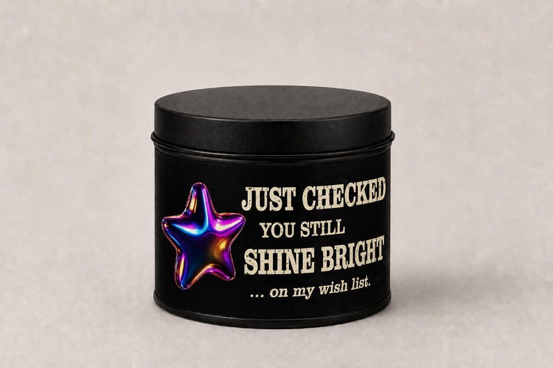 Puede incluir: Una lata de vela redonda negra con tapa. La lata presenta una estrella hologr&aacute;fica y el texto "JUST CHECKED YOU STILL SHINE BRIGHT ... on my wish list."