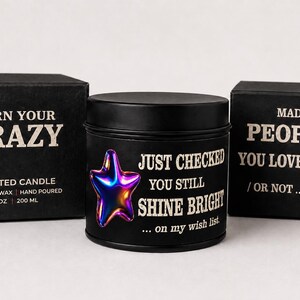 Puede incluir: Vela perfumada negra con un dise&ntilde;o de estrella met&aacute;lica y el texto "Just Checked You Still Shine Bright". La vela est&aacute; en una lata negra, con una caja a juego que dice "Burn Your Crazy". La otra caja dice "Made for People You Love".