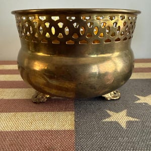 May include: A polished brass bowl featuring a rounded body and a decorative openwork lattice rim. The bowl is supported by three small, ornate feet, providing a stable base. The metal surface exhibits a warm, metallic gold tone with subtle reflections.