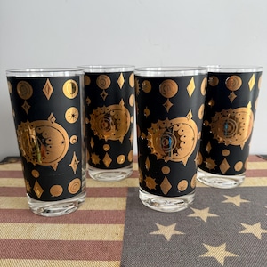 May include: A set of four cylindrical drinking glasses featuring a matte black exterior finish with metallic gold celestial patterns, including suns, moons, stars, and diamond shapes arranged in a repeating motif around the glass surface.