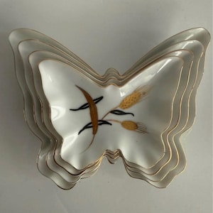 May include: A set of three nested white porcelain dishes shaped like butterflies. Each dish features a gold-trimmed scalloped edge and a central graphic of golden wheat stalks with black stems. The dishes are stacked to show their wing-like forms.