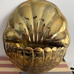May include: A polished brass wall pocket shaped like a large scallop shell. The metal surface features a reflective gold finish with vertical fluted ridges and a decorative beaded edge along the curved top border of the shell-shaped container.