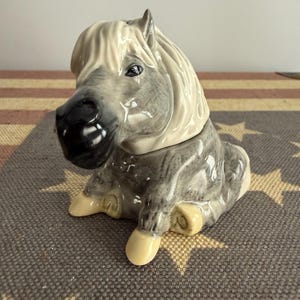 May include: A ceramic horse figurine featuring a grey coat with a dark muzzle and a cream-colored mane. The horse is depicted in a seated position with its front legs extended forward, showcasing detailed sculpting and a glossy finish throughout.