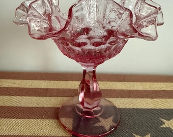 May include: A translucent pink glass compote dish featuring a wide, ruffled, and crimped rim. The bowl has a textured hobnail pattern on the exterior surface, supported by a thick, faceted pedestal stem and a circular, flat glass base.