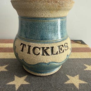 May include: A ceramic jar with a lid, featuring a speckled beige finish and a blue glazed band. The word TICKLES is embossed in dark capital letters on the front. The jar has a flared rim and a rounded body with a scalloped blue glazed base.