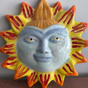 May include: A ceramic sun wall plaque featuring a light blue face with a tan forehead section. The sun has yellow rays radiating outward, each accented with vertical red painted stripes. The facial features include almond-shaped eyes and lips.