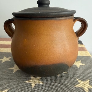 May include: A round, two-handled ceramic pot with a matching lid. The pot features a warm, earthy brown body that transitions into a dark, charcoal-black finish at the base and rim. The lid is a solid dark charcoal-black with a central knob.