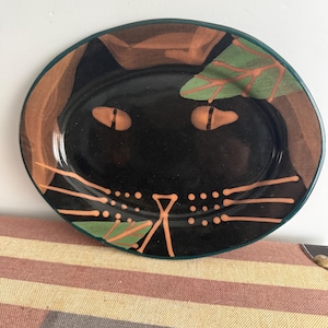 May include: An oval ceramic plate featuring a glossy black cat face design. The cat has orange almond-shaped eyes, a triangular nose, and long whiskers. Green leaves are painted in the upper right and lower left corners.