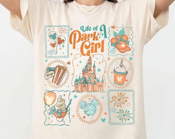 May include: Ivory-colored t-shirt with a graphic design featuring the text "Life of a Park Girl" and various illustrations. The design includes a castle, popcorn, and other park-themed images. The shirt is labeled "Comfort Colors Ivory".