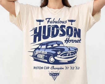 May include: Ivory-colored t-shirt featuring a blue graphic of the Hudson Hornet race car with the text "Fabulous Hudson Hornet" and "Piston Cup Champion '51 '52 '53". The shirt is part of the Comfort Colors collection.